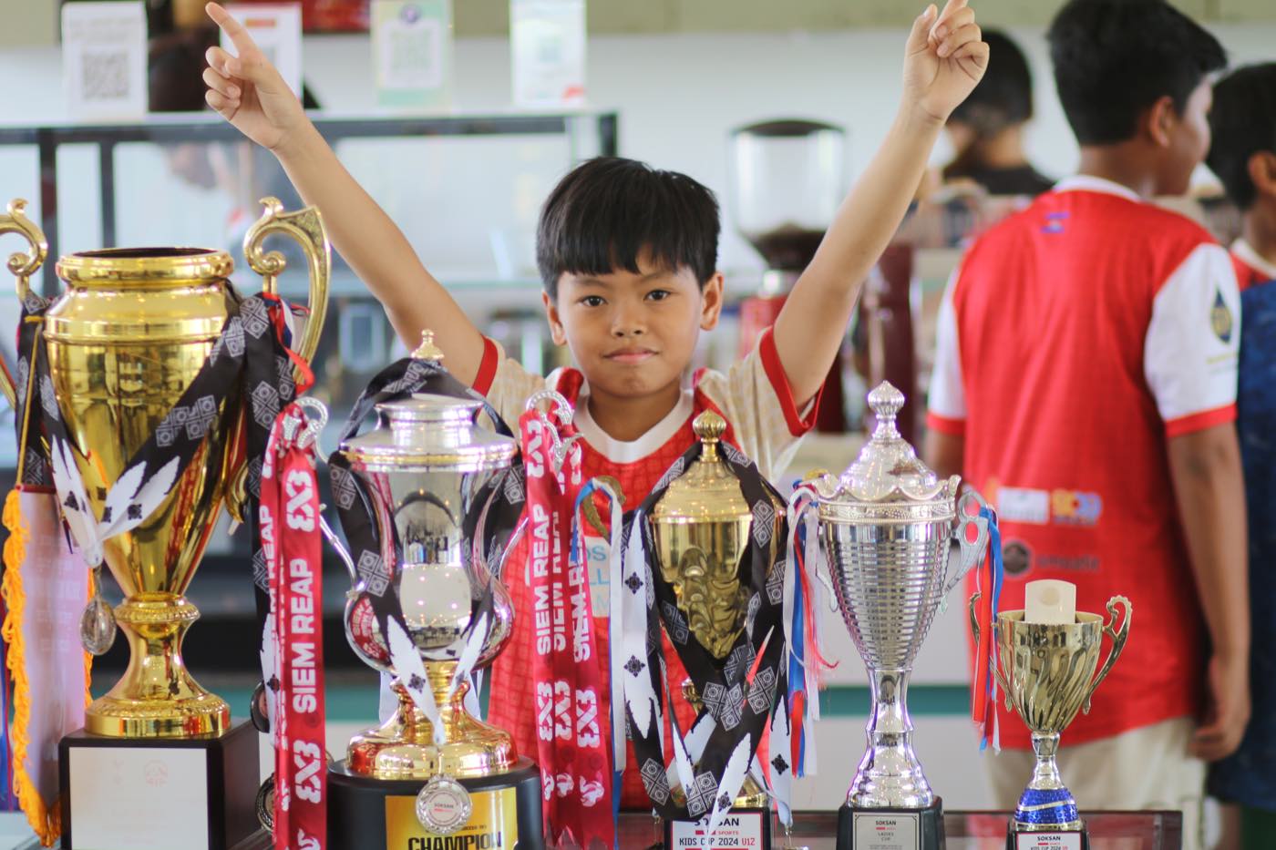 A young player raising both arms behind a table of tournament trophies