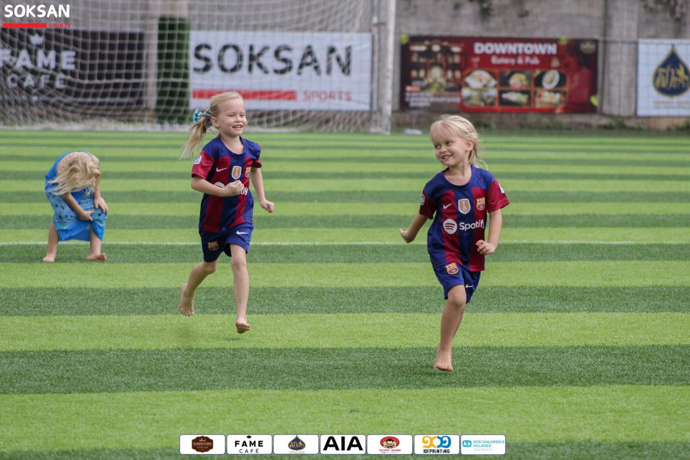 Two small girls in football kit running barefoot across the pitch past the SOKSAN banner